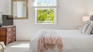 A bedroom with a bed, a dresser, a television, a window with a view of greenery, and a mirror on the wall. - Pip's Cottage - Pets Friendly, Cafes, Bars, Jetty Beach (Coffs Harbour)