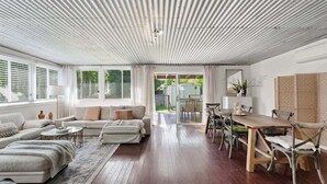 A spacious living area with a wooden dining table, cushioned seating, and a view through a glass door to an outdoor space. - Pip's Cottage - Pets Friendly, Cafes, Bars, Jetty Beach (Coffs Harbour)