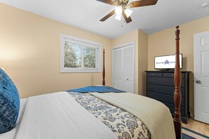 Une chambre à coucher avec un lit à baldaquin, un ventilateur de plafond, un téléviseur posé sur une commode et une fenêtre avec des stores.