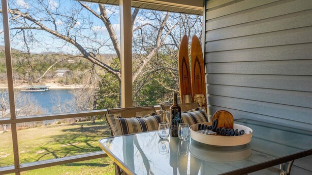 Een glazen tafel met een schaal, glazen en een fles op een veranda met uitzicht op een meer en bomen.