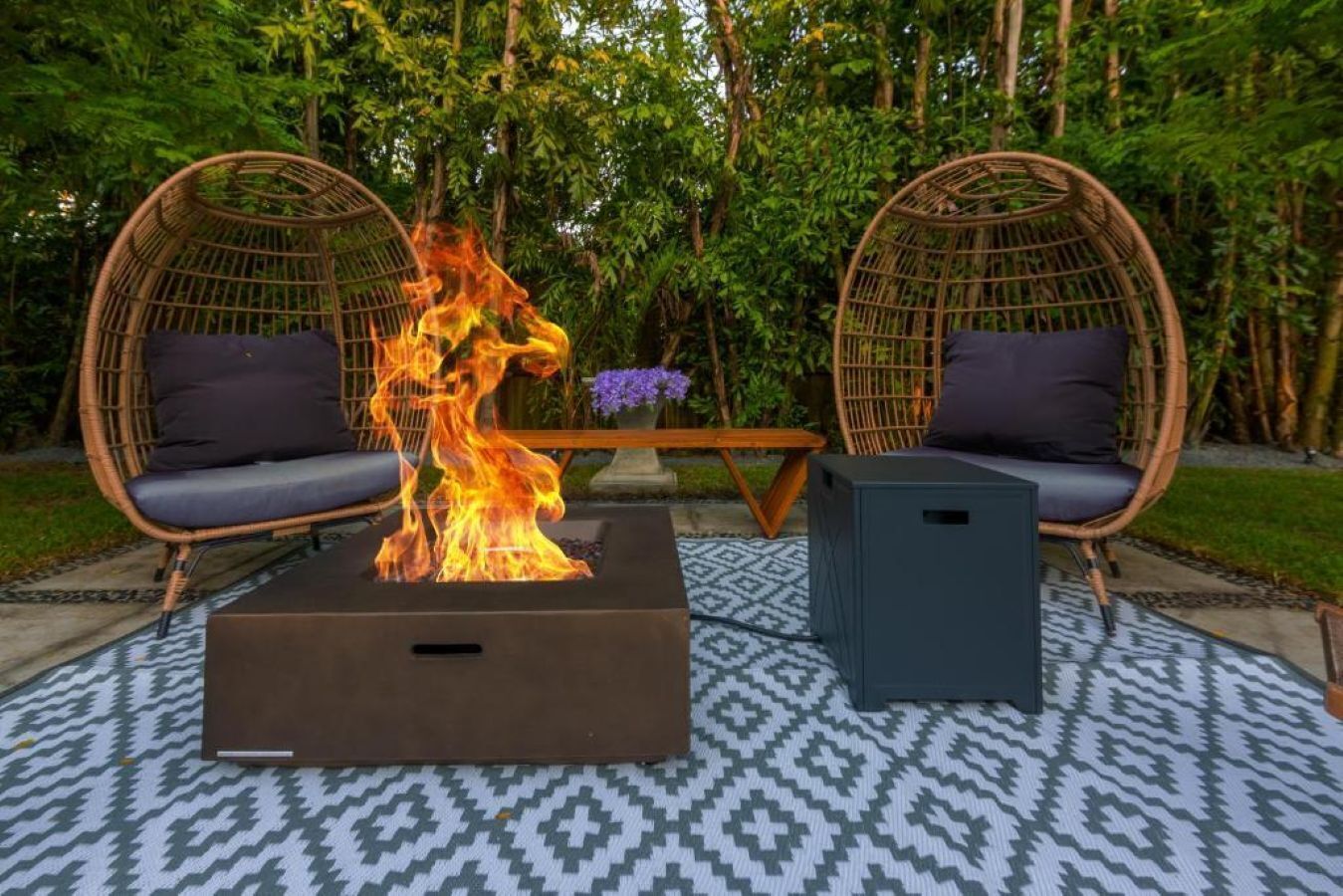 Un foyer au centre, deux fauteuils en osier avec des coussins, une petite table et un objet rectangulaire de couleur sombre.