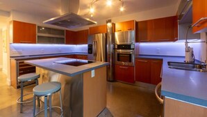 A modern kitchen with stainless steel appliances, wooden cabinets, and a central island with stools. - Villa II Palazzo by Glam Homes (North Miami)