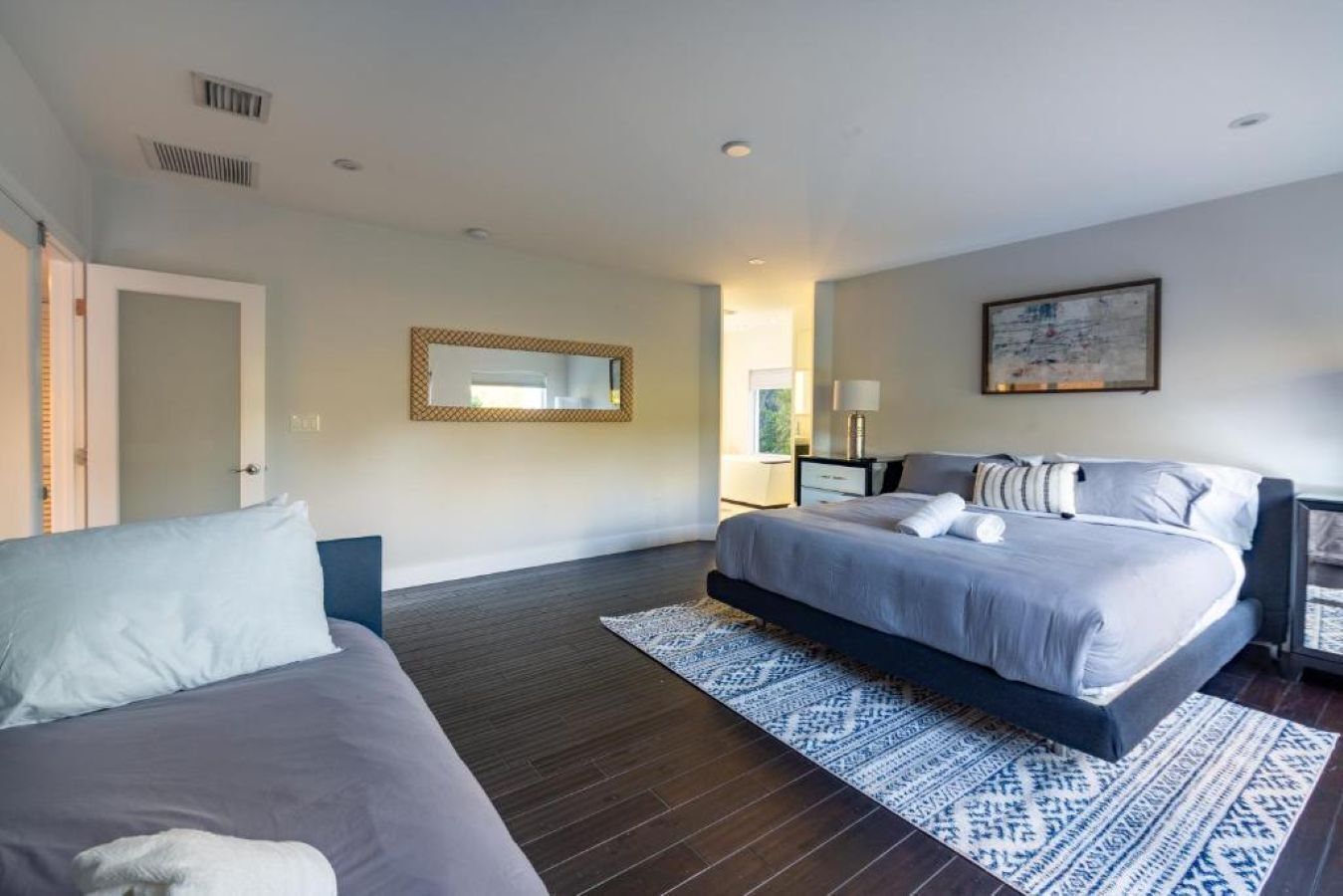 A modern bedroom with a large bed, a smaller sofa, a patterned rug, a mirror, and a painting on the wall.