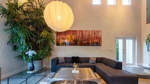 A modern living room with a sectional sofa, a glass coffee table, a large potted plant, and a wall-mounted painting. - Villa II Palazzo by Glam Homes (North Miami)