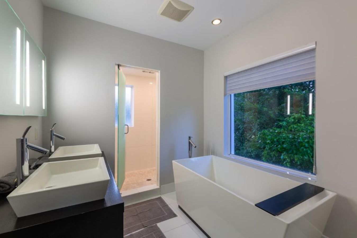 Ein modernes Badezimmer mit zwei Waschbecken, einer Badewanne und einem Fenster mit Blick ins Grüne.