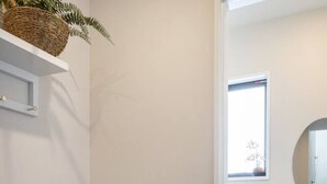Un pasillo con una planta en maceta, un estante blanco y una puerta que da a un baño con inodoro y espejo.