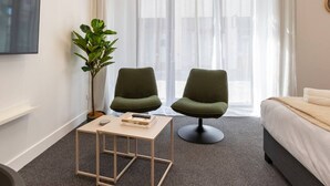 A hotel room with a bed, two green armchairs, a small table with books, a plant, and a TV on the wall. - Hotel Comfort | Ground Floor, City Apartment (Christchurch)