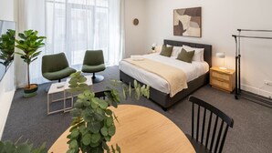 A modern hotel room with a bed, two green chairs, a round table, and a potted plant. - Hotel Comfort | Ground Floor, City Apartment (Christchurch)