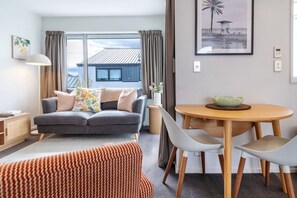A modern living room with a grey sofa, a dining table with chairs, a patterned ottoman, and a large window with curtains. - City Fringe Stay + Garage & Sunny Balcony (Christchurch)