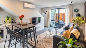 A modern living room with a sofa, a dining table with chairs, a TV, and a large window. - Stadium Base + Sunny Courtyard Living (Christchurch)