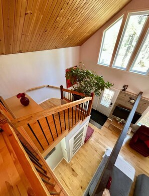 A loft-style room with a wooden ceiling, a dining area with a table and chairs, a large window, and a staircase.