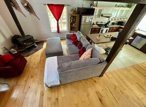 Una sala de estar con un sofá, un sillón rojo, piso de madera, una cocina al fondo y una cortina roja.