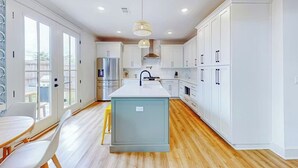 A modern kitchen with white cabinetry, a central island, and a dining area with a table and chairs. - Perdido Bayside Retreat Near Beach! Pool, Dock, Pickleball (Pensacola)