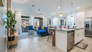A modern kitchen with a central island, white cabinetry, stainless steel appliances, and a dining area with blue furniture. - 6BR/4.5BA Luxury Disney Decor w/ Pvt Pool & Resort (Kissimmee)