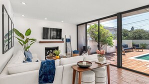 A modern living room with a fireplace, a flat-screen TV, and a large sliding glass door leading to a patio with outdoor furniture. - New! La Jolla Shores 2BD with Pool- Walk to Beach! (La Jolla)