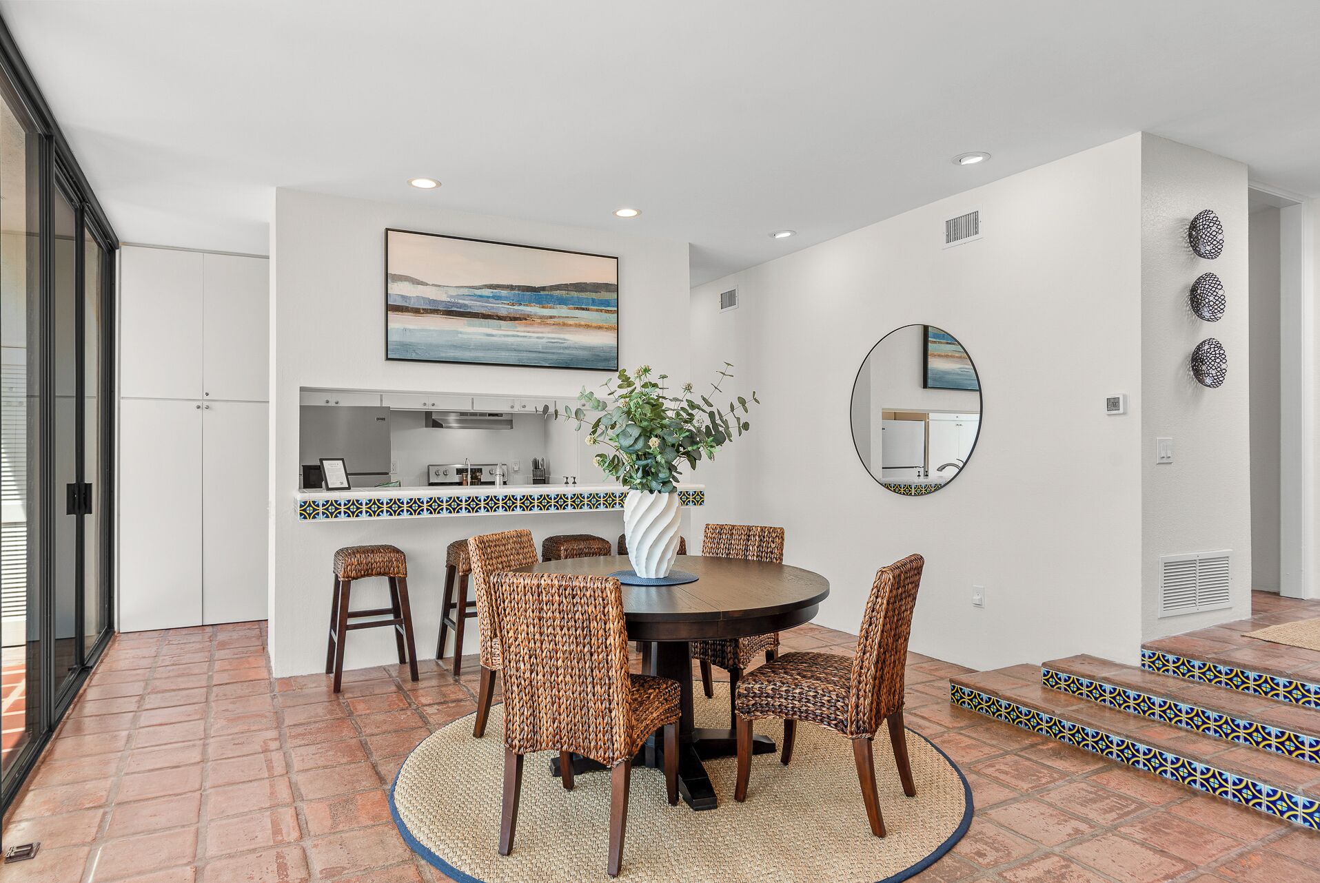 A dining area with a round table, wicker chairs, a vase with greenery, a kitchen in the background, and a large painting on the wall.