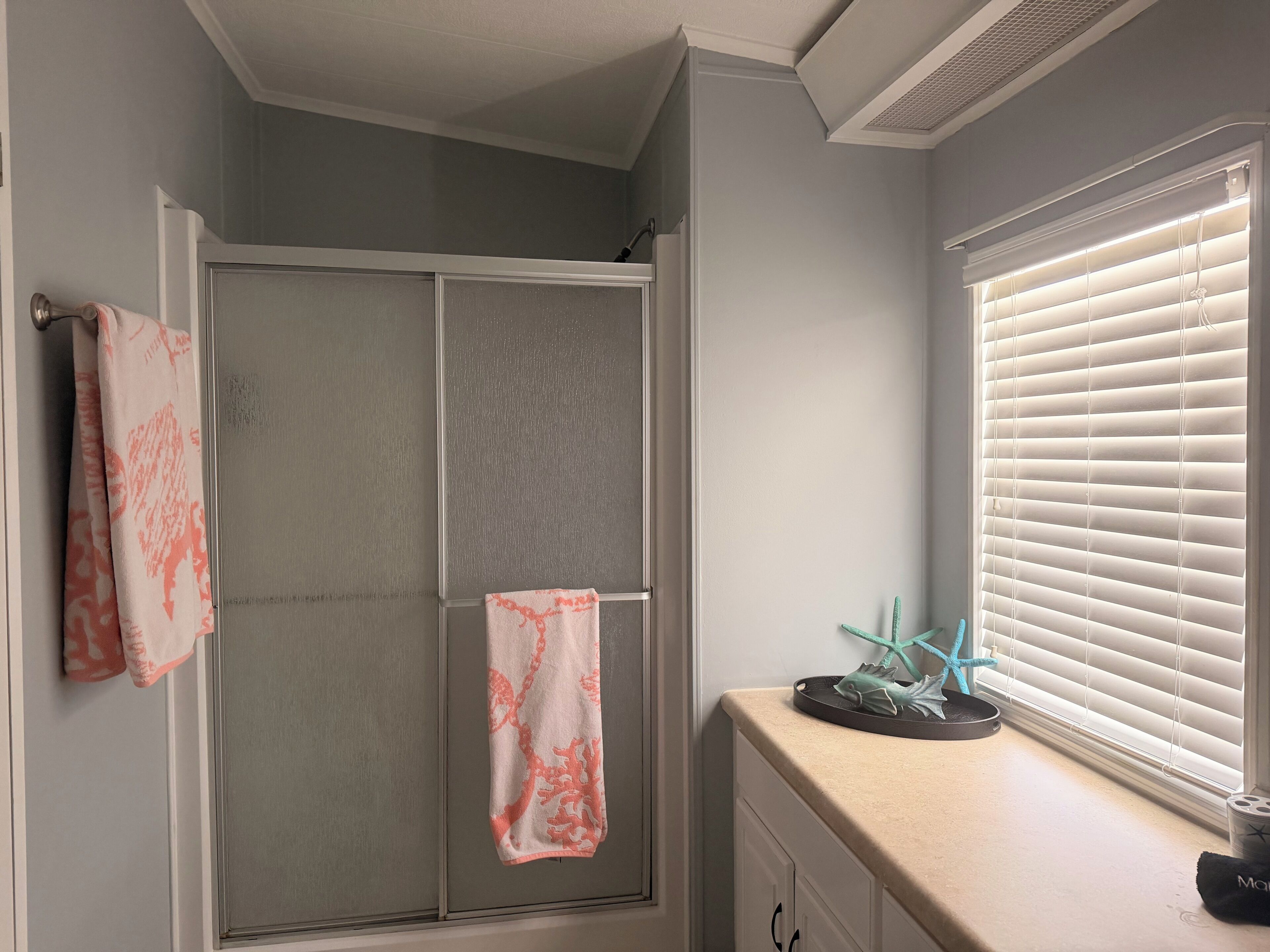 A bathroom with a shower door, a towel rack with a towel, a window with blinds, and a small shelf with a decorative item.