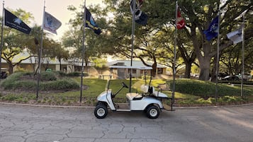 Un carrito de golf estacionado en un estacionamiento con varias banderas, árboles y un edificio al fondo.