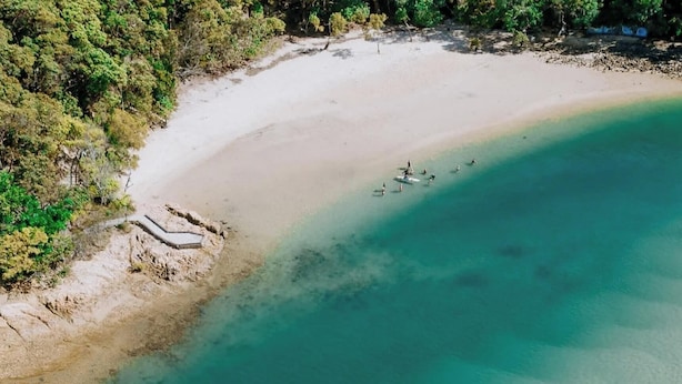 Eine Vogelperspektive zeigt einen Strand mit klarem türkisblauem Wasser und einem sandigen Ufer, auf dem sich einige Personen im Wasser befinden.