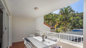 Ein Balkon mit weißem Tisch und Stühlen, einem weißen Geländer und Blick auf Palmen sowie ein blaues Schwimmbecken.