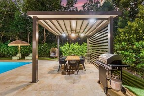 Un patio al aire libre con una parrilla, una mesa, sillas y una pérgola.
