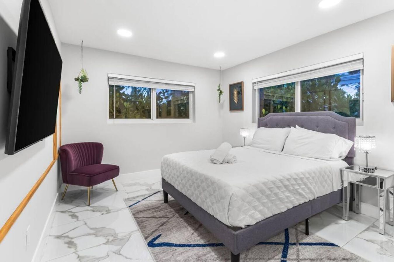 A modern bedroom with a bed, a TV, a chair, and two windows.
