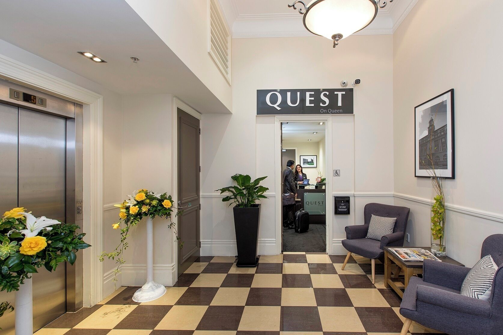 A hotel lobby with a checkered floor, a reception desk, a sign reading 'QUEST On Queen,' and a reception area.