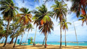 A beach with palm trees, a lifeguard tower, and the ocean in the background. - Elevate Your Stay! Prime 2 Beds Home w Ocean View! (San Juan)