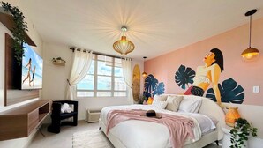 A bedroom with a bed, a TV, a window with curtains, a wall mural, and a ceiling light fixture. - Elevate Your Stay! Prime 2 Beds Home w Ocean View! (San Juan)