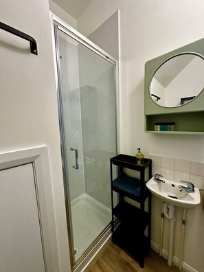 A bathroom with a glass shower enclosure, a sink with a mirror above it, and a small shelf with toiletries. - The Joseph Cosy at Lancaster (Lancaster)