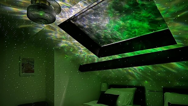 Une pièce comprenant un lit, un luminaire suspendu et une projection sur le mur représentant une nébuleuse verte.