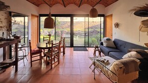 A spacious room with a brick ceiling, a dining area with a table and chairs, a sofa, and large sliding glass doors leading to an outdoor area with rocks and greenery. - Casa Tacuba country house near the mountain and lake Chapala (San Juan Cosalá)