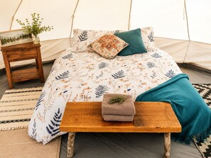 A tented room with a bed, a small wooden table with a towel, a vase with greenery, and a picture frame on a nightstand.