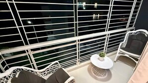 Ein Balkon mit weißen Metallgeländern, einem runden Tisch mit einem Blumentopf und zwei weißen Korbstühlen mit Kissen.