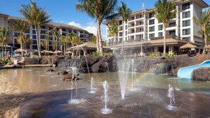 Un hotel con una fuente ornamental y un tobogán, rodeado de palmeras y una piscina.