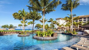 Zona de piscina en un resort, con palmeras, sillones de descanso y un edificio al fondo.