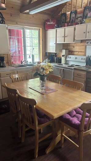 Una mesa de comedor de madera con sillas, un florero con flores, una cocina con armarios blancos, una estufa y una ventana con vista a follaje.