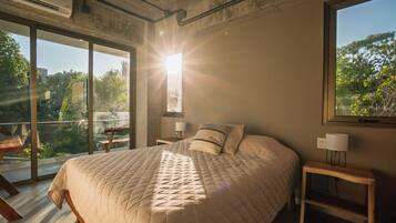 Un dormitorio con una cama grande, dos mesitas de noche con lámparas, vistas a zonas verdes a través de amplios ventanales y el sol brillando en la ventana.
