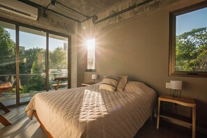 Un dormitorio con una cama grande, dos mesitas de noche con lámparas, vistas a zonas verdes a través de amplios ventanales y el sol brillando en la ventana.