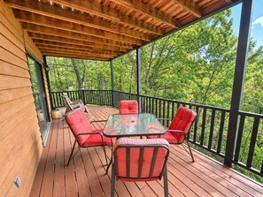 A covered deck with a glass table and red chairs, surrounded by a black railing, overlooking a forested area. - Mountain Ridge-VIEWS-Pigeon Forge | Hot Tub, Decks & Fireplace | Sleeps 4 (Pigeon Forge)