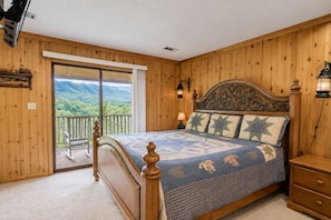 A bedroom with a wooden headboard, a bed with patterned bedding, a nightstand, a lamp, and a view of mountains through a sliding door. - Mountain Ridge-VIEWS-Pigeon Forge | Hot Tub, Decks & Fireplace | Sleeps 4 (Pigeon Forge)