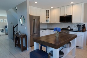 A modern kitchen with white cabinetry, a stainless steel refrigerator, a wooden island, and a vase with blue flowers. - Sun Drenched, 0.1 mi to Bike Path, <1 mi to Beaches, Peloton, Games (Chatham)