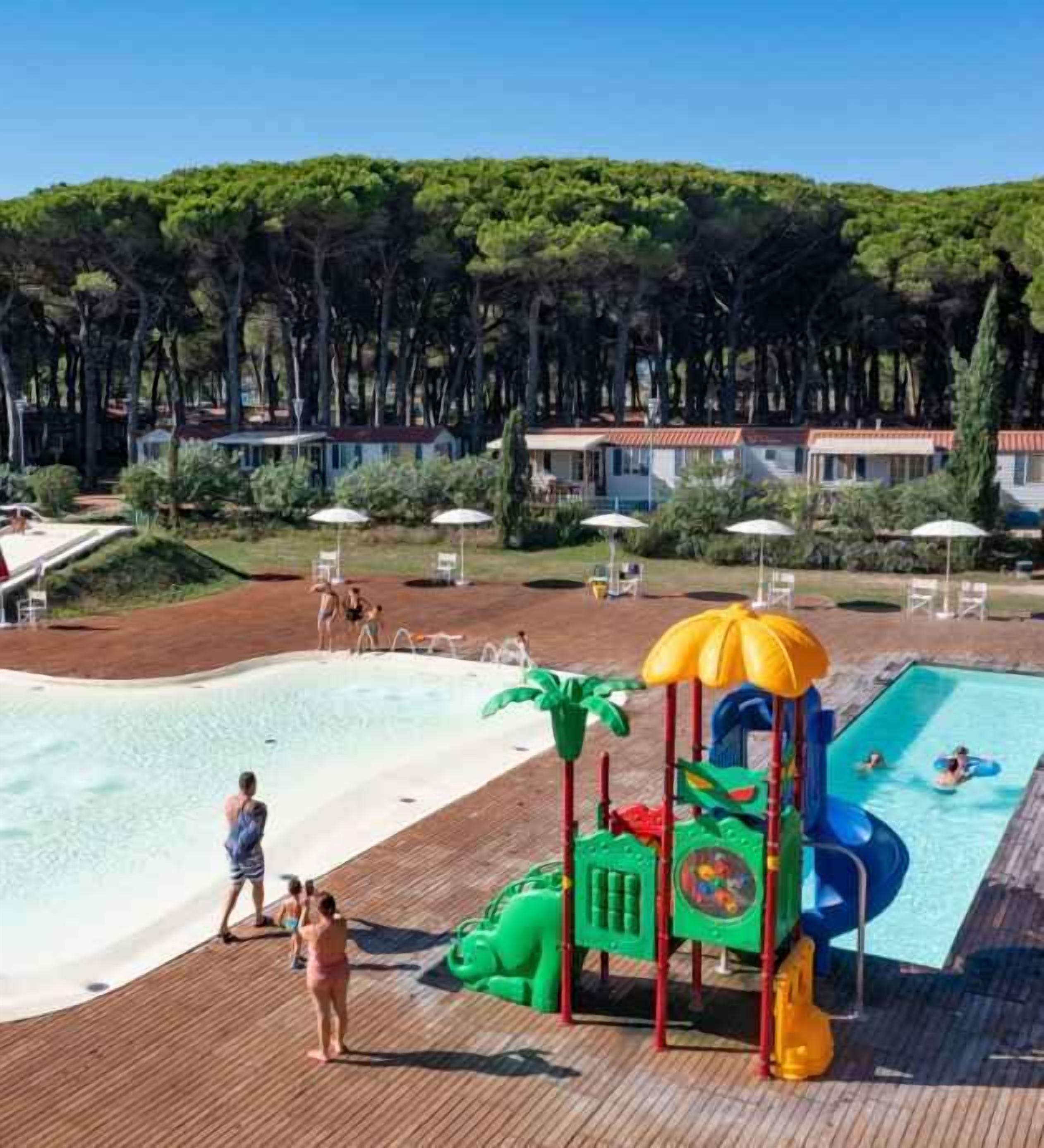 Zona de piscina amb tobogan aquàtic, zona de jocs i vista a una zona boscosa.