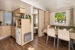 Une cuisine compacte équipée d’un four à micro-ondes, d’un réfrigérateur et de meubles de rangement en bois. On y trouve également un coin repas avec une table et des chaises, ainsi qu’une fenêtre donnant sur un espace verdoyant.