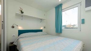 Een slaapkamer met een bed, een raam met gordijnen, een wandplank met een plant en een nachtkastje.