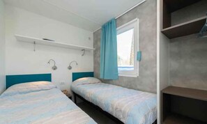 Een kleine kamer met twee bedden, een raam met gordijnen, een plank boven de bedden en een wandlamp.