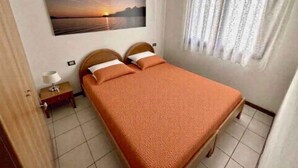 Ein Einzelbett mit Kopfteil, ein Nachttisch mit Lampe, ein Fenster mit durchsichtigen Vorhängen und ein Sonnenuntergangsbild an der Wand.
