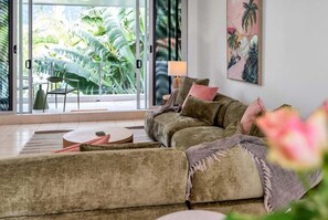 A living room with a sofa, a round coffee table, and a view of tropical foliage through large glass doors. - Belle Escapes Tides & Palms (Palm Cove)