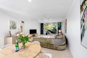 A modern living room with a sofa, a coffee table, a TV, and a dining area with a table and chairs. - Belle Escapes Tides & Palms (Palm Cove)