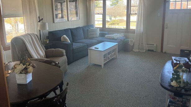 A living room with a sofa, a coffee table, a dining table with a vase of flowers, and large windows with curtains.